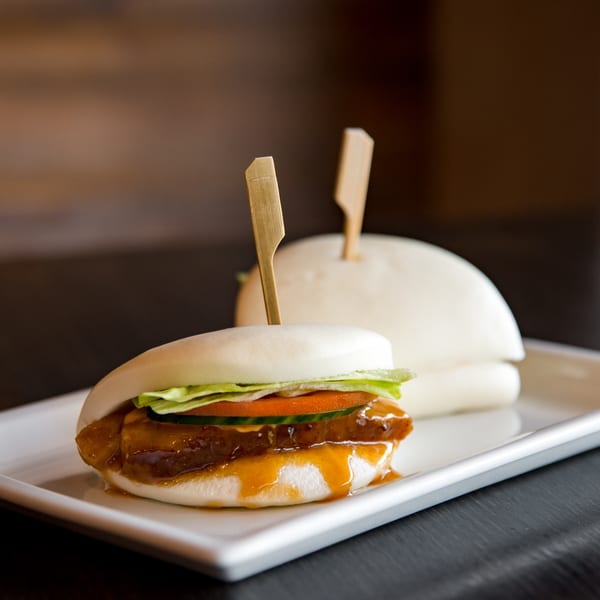 Two bao buns with pork belly, lettuce, and cucumber on a white rectangular plate.