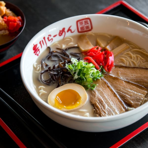 A bowl of ramen with pork, egg, and vegetables, with a side dish of chicken and rice.