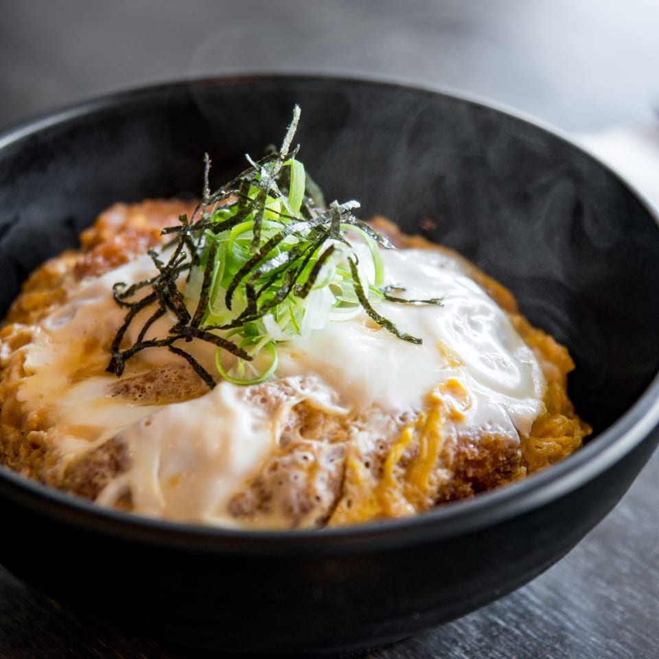 Steaming katsudon with a fried egg, green onions, and seaweed in a black bowl.