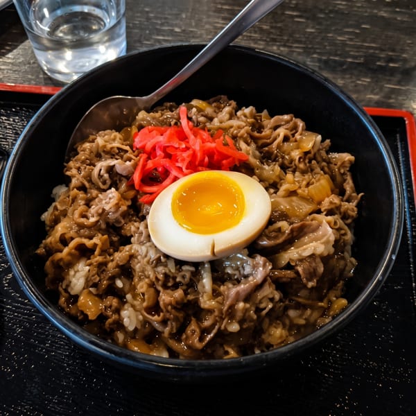 A bowl of gyudon with thinly sliced beef, onions, red ginger, and a soft-boiled egg.
