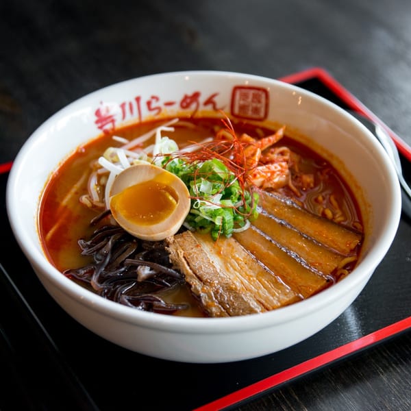 Spicy ramen with pork belly, soft-boiled egg, bean sprouts, and scallions in a white bowl.